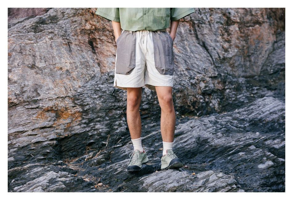 Front view of a model wearing ivory shorts with large gray front pockets, standing on rocky terrain.