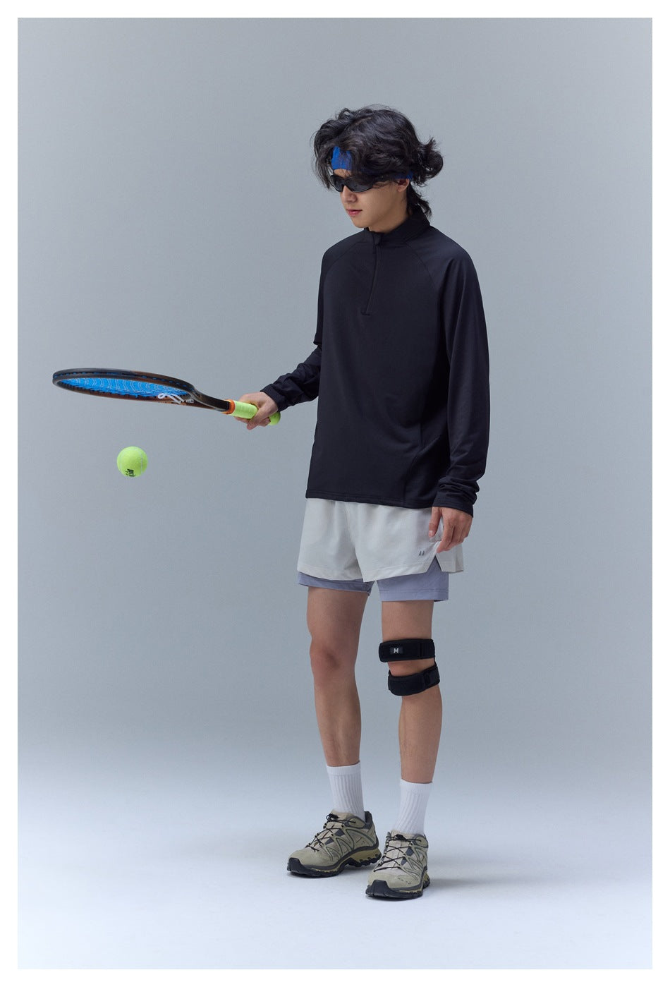 Model wearing a black long-sleeve quarter-zip top with white and lavender shorts while holding a tennis racket.
