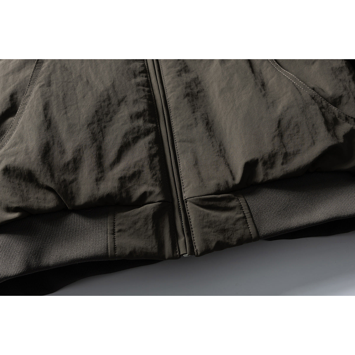 Close-up of an olive-gray jacket's front zipper, lower hem, and rib-knit waistband.