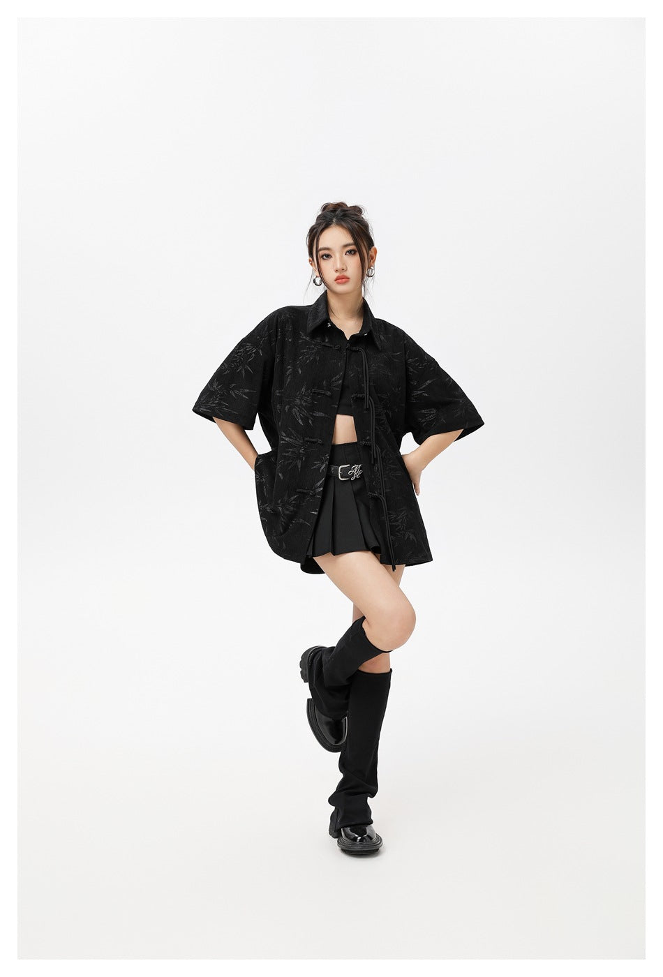 Model wearing a black patterned short-sleeve shirt with tie closures over a black crop top and pleated skirt.