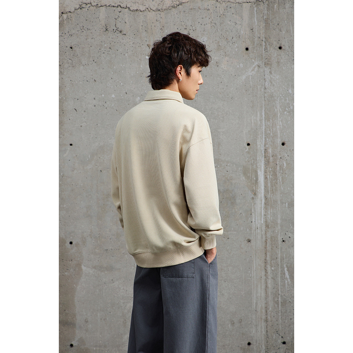 Model seen from behind wearing a light beige collared pullover with ribbed cuffs and hem, paired with gray trousers.