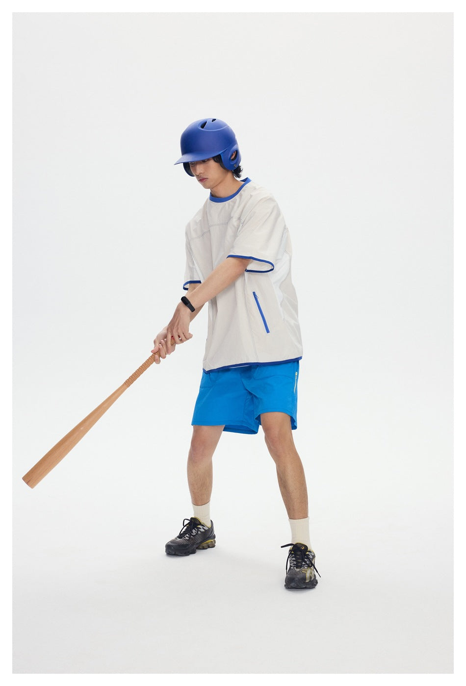 Model in a side pose wearing an ivory shirt with blue trim and bright blue shorts, holding a bat.