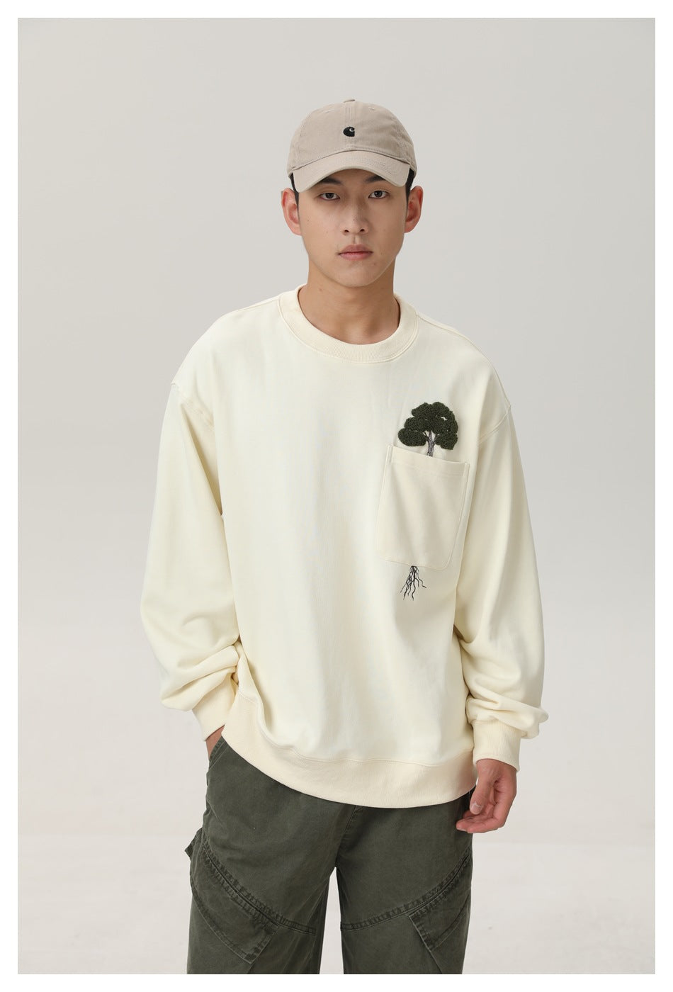 Model wearing an ivory crewneck sweatshirt with a chest pocket and embroidered tree, paired with olive pants and a beige cap.