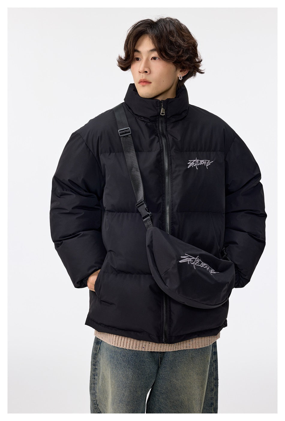 Model wearing a black high-collar puffer jacket with a small matching crossbody bag and white script details.