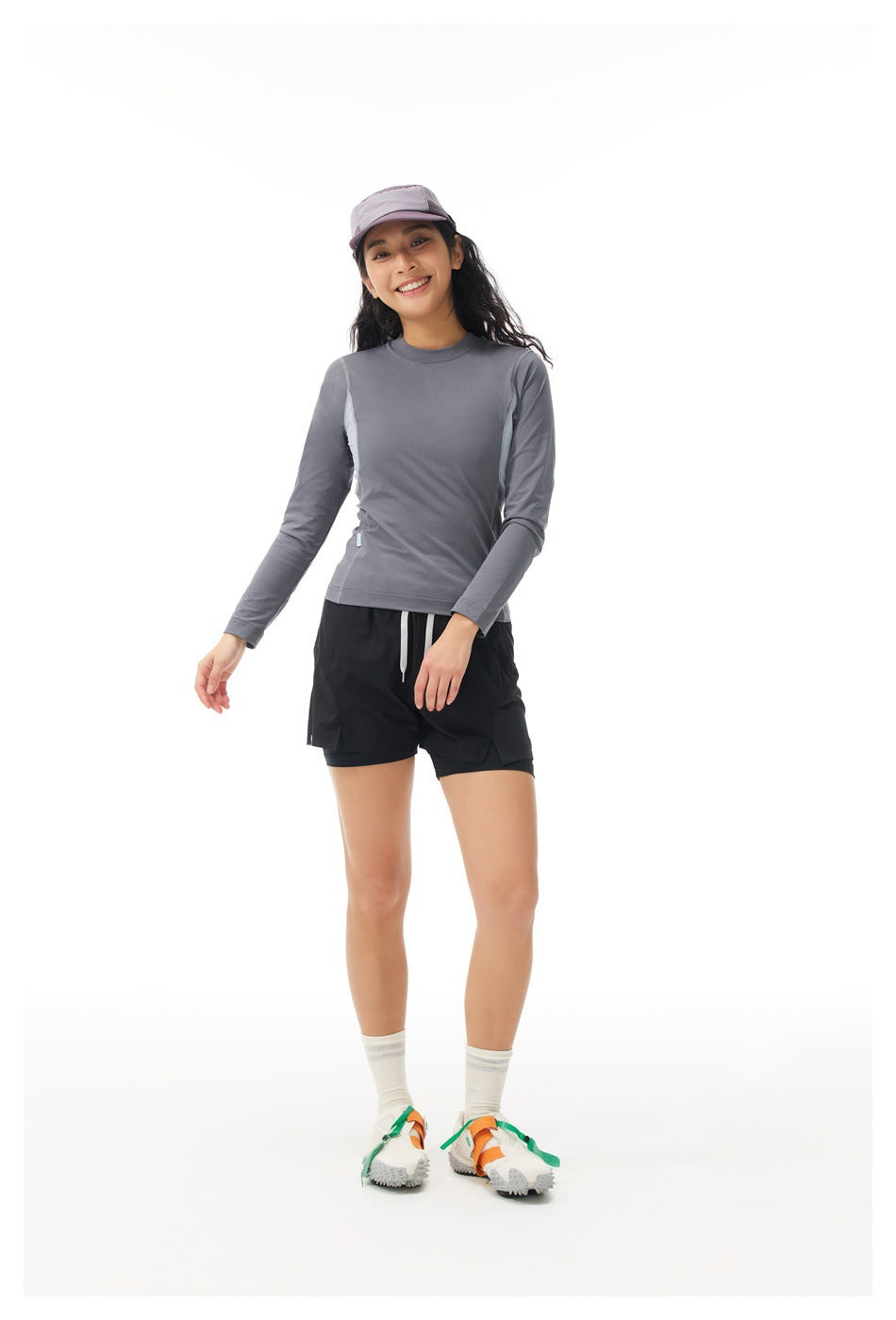 Full-length model wearing a gray long-sleeve top, black shorts, a lavender cap, white socks, and colorful shoes.