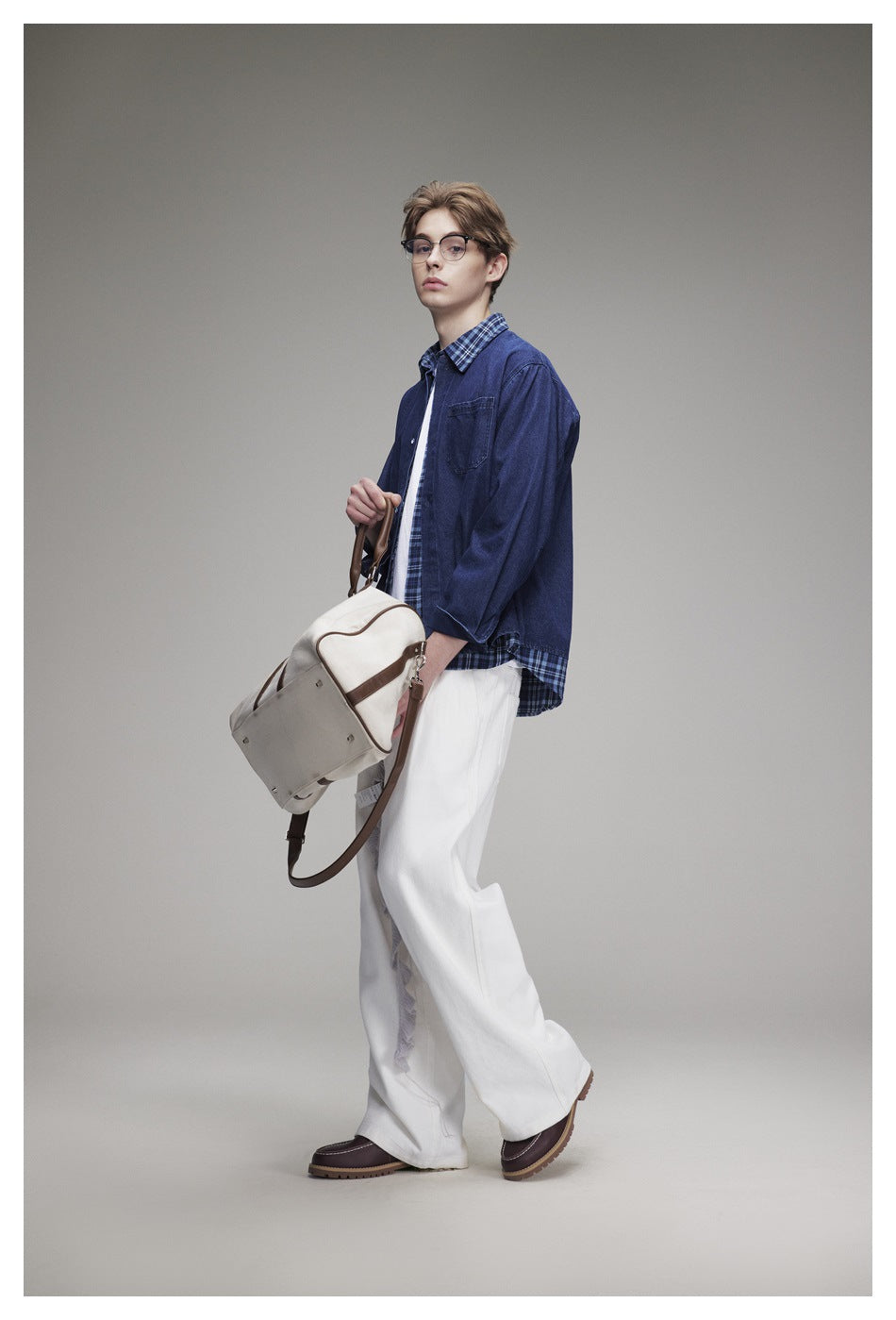 Model wearing a blue denim overshirt with plaid trim, white wide-leg trousers and brown shoes, carrying a cream bag.