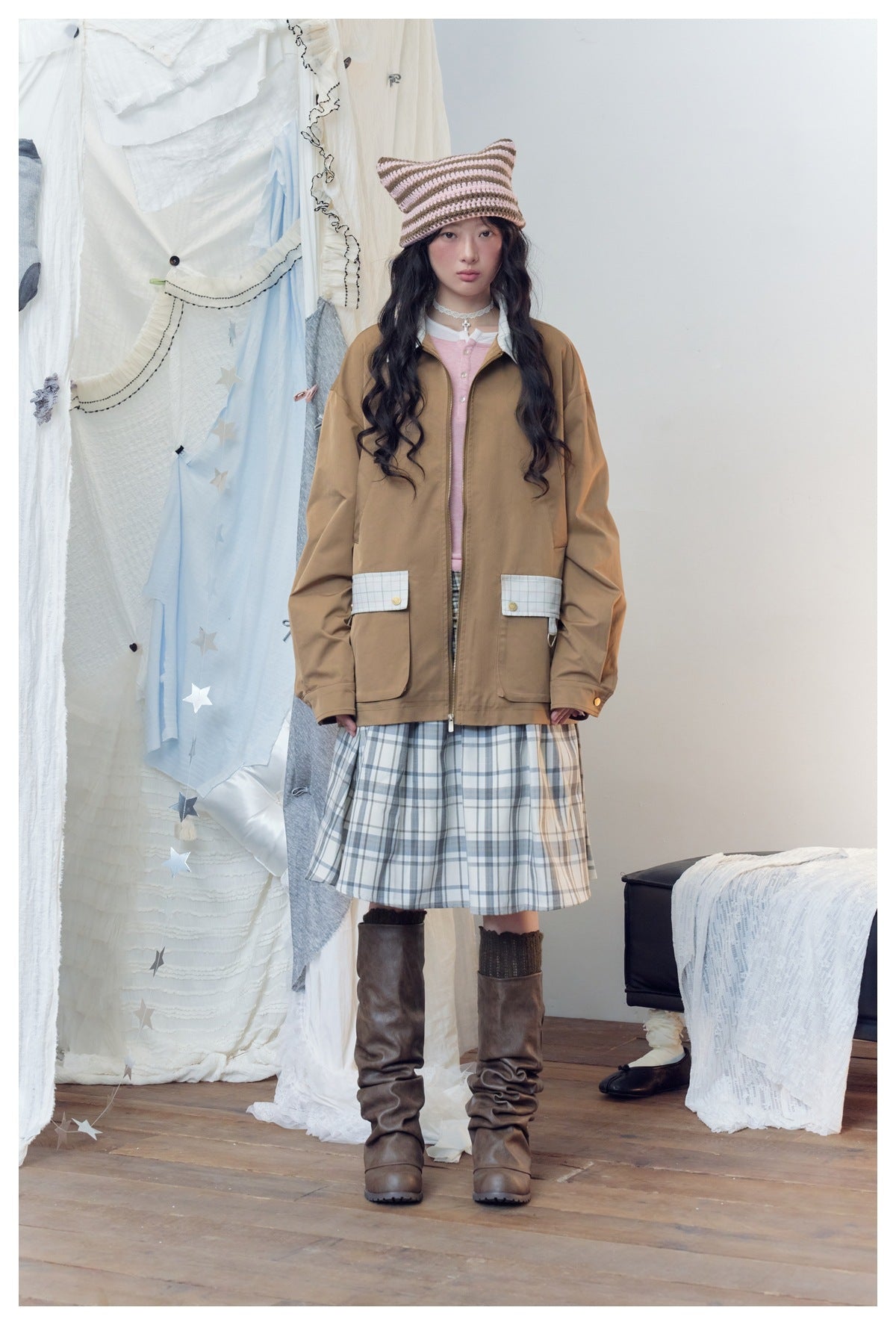 Model wearing the khaki jacket zipped over a pink top and plaid skirt, front view, with a striped beanie and boots