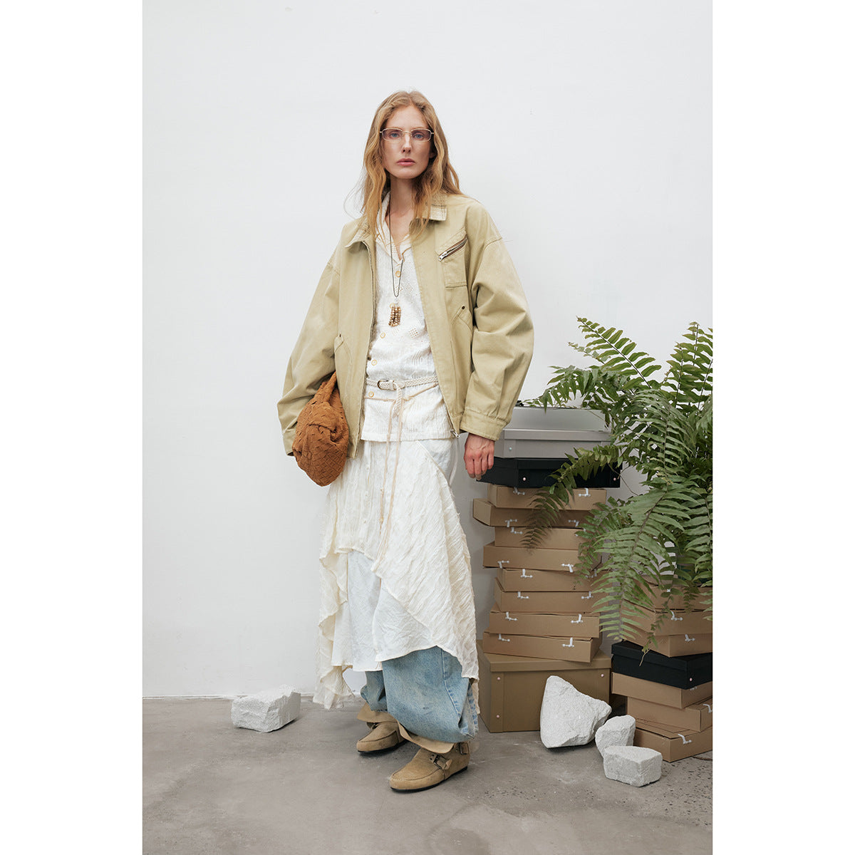 Full-length view of a model wearing a loose beige jacket over a layered white outfit and blue jeans.