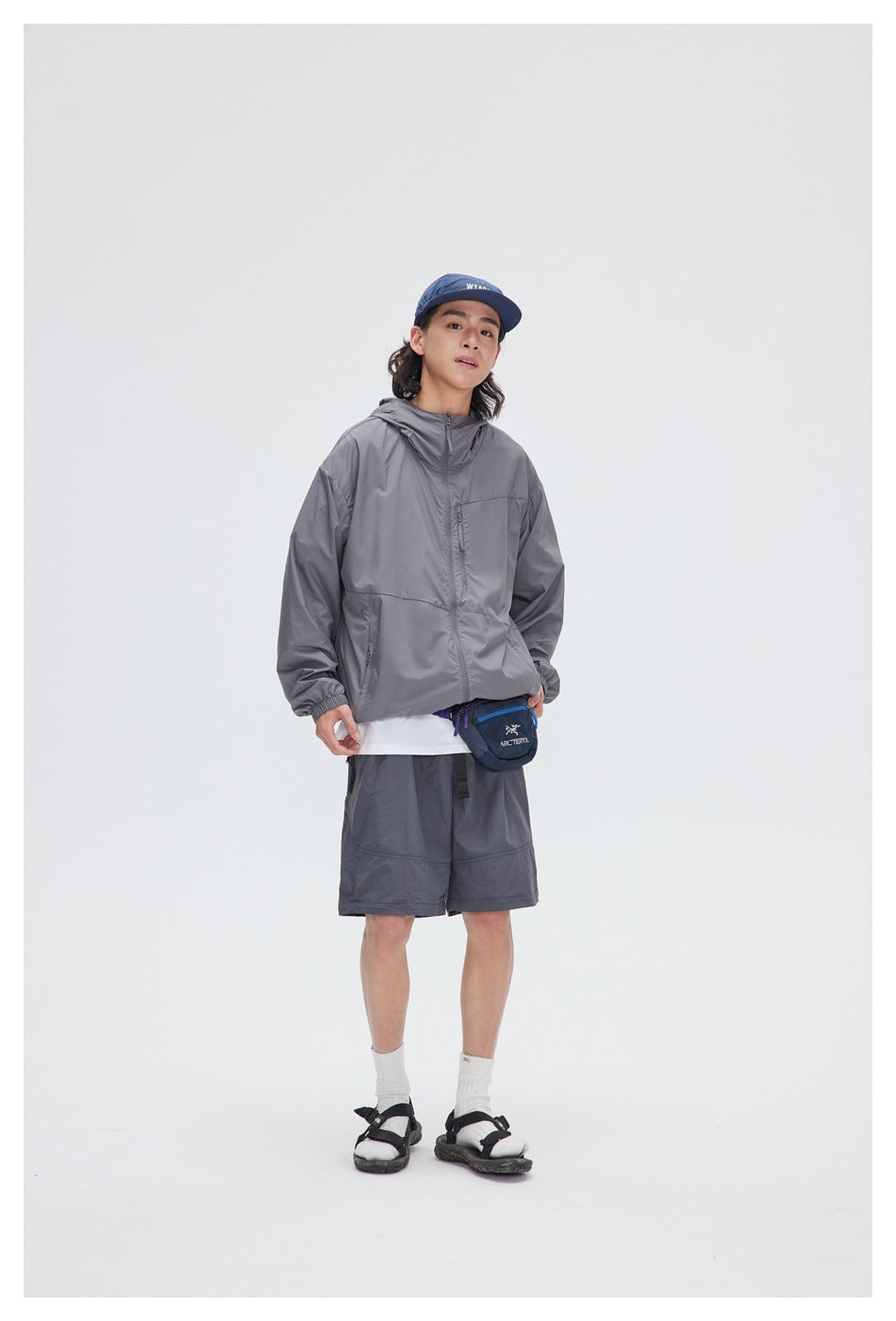 Person wearing a gray hooded zip jacket with gray shorts, blue cap, white socks and sandals