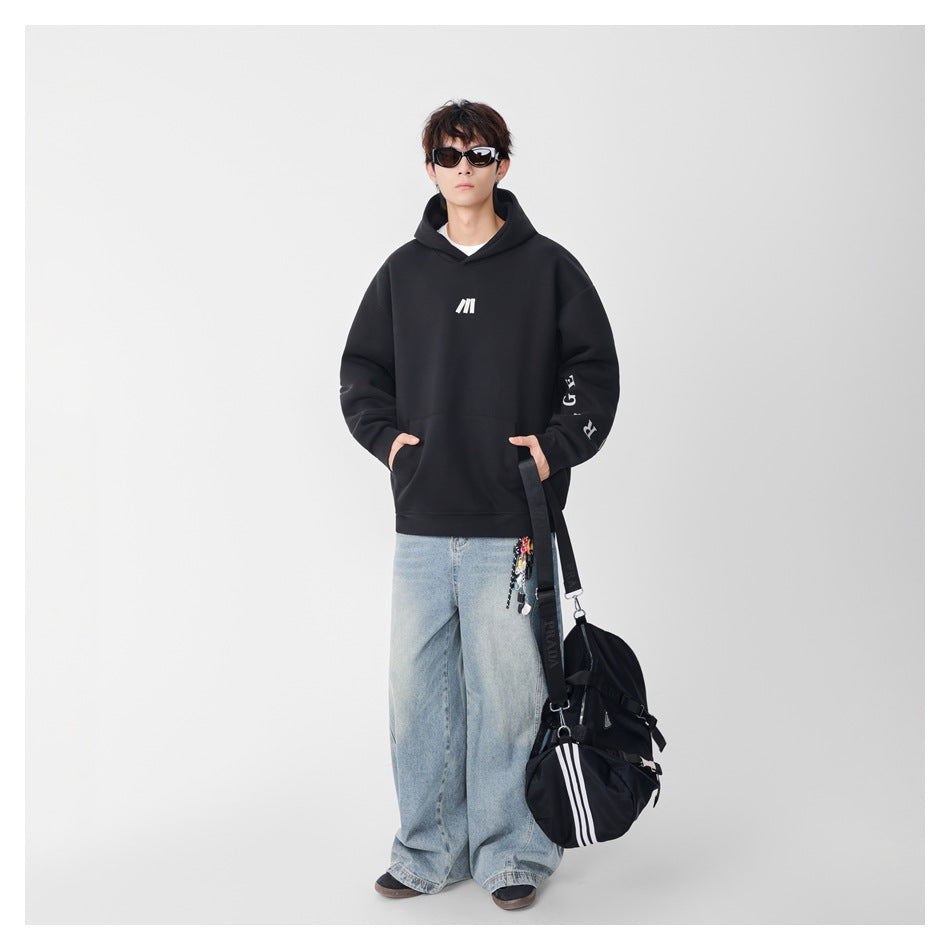 Model wearing a black oversized hoodie with a small white chest mark and sleeve lettering, paired with light blue jeans.