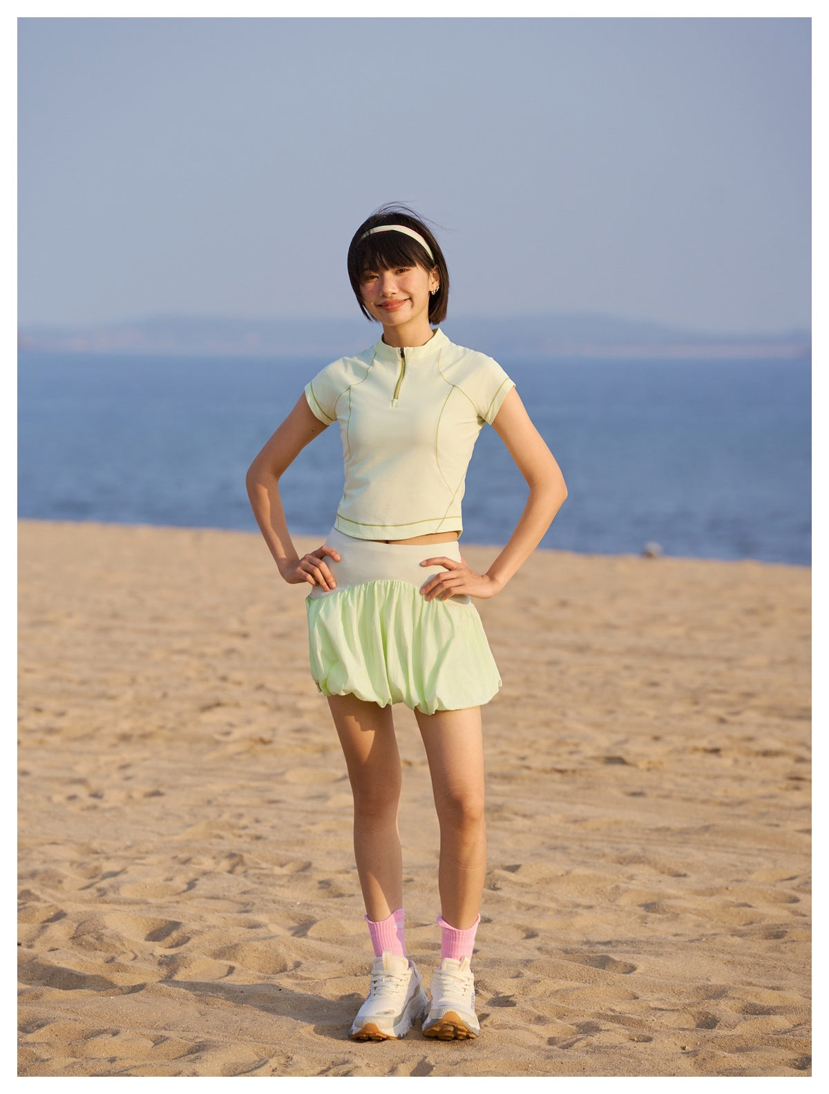 Model wearing a pale yellow-green zip-neck top and bubble skirt on a beach, shown full length with pink socks and sneakers.