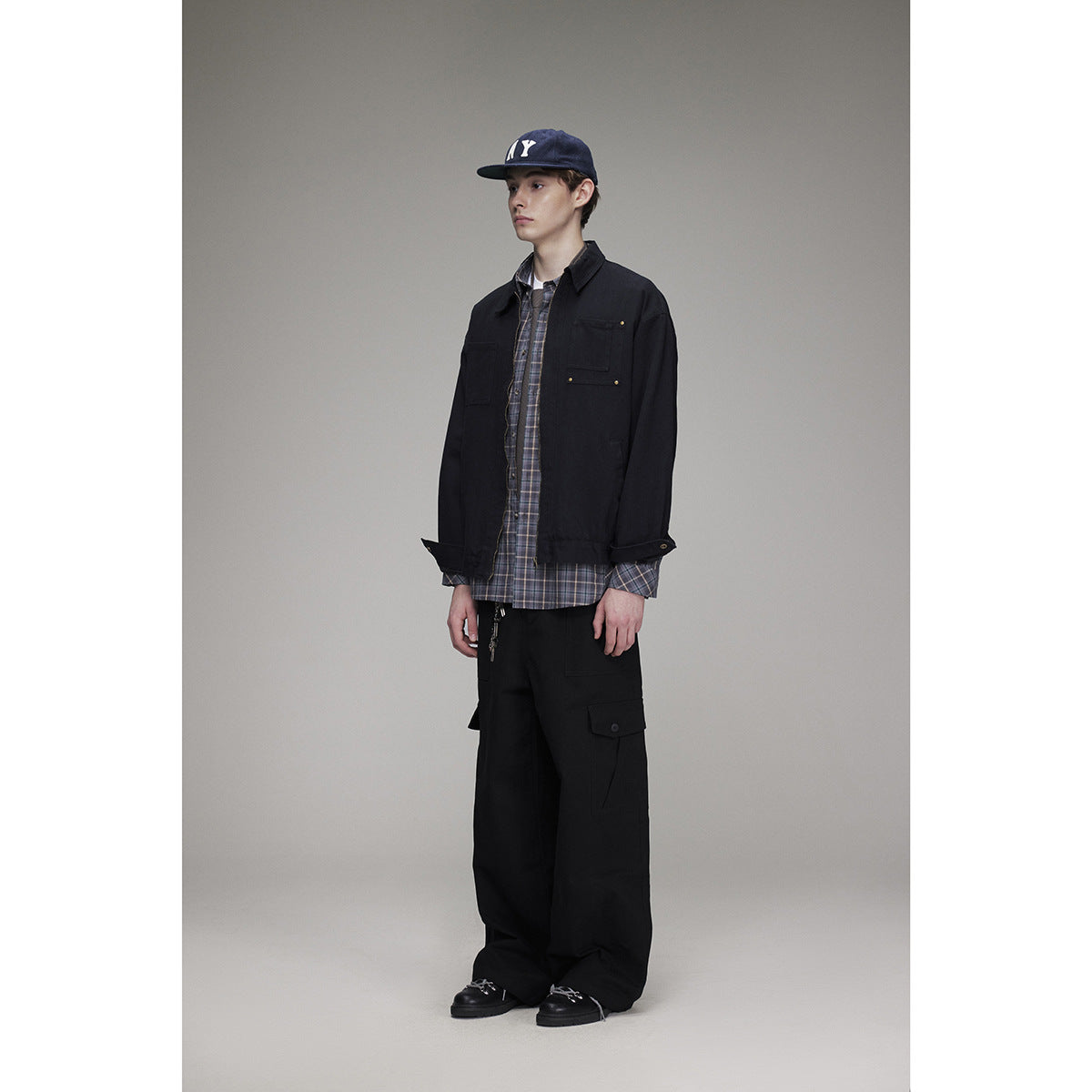 Model in a black utility jacket and cargo pants, layered over a plaid shirt, wearing a navy cap.