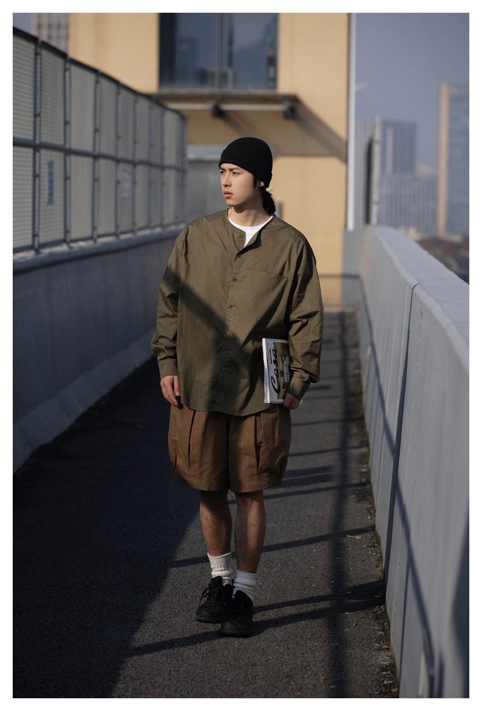 Model wearing an olive button-front shirt and loose brown shorts, shown outdoors from the front.