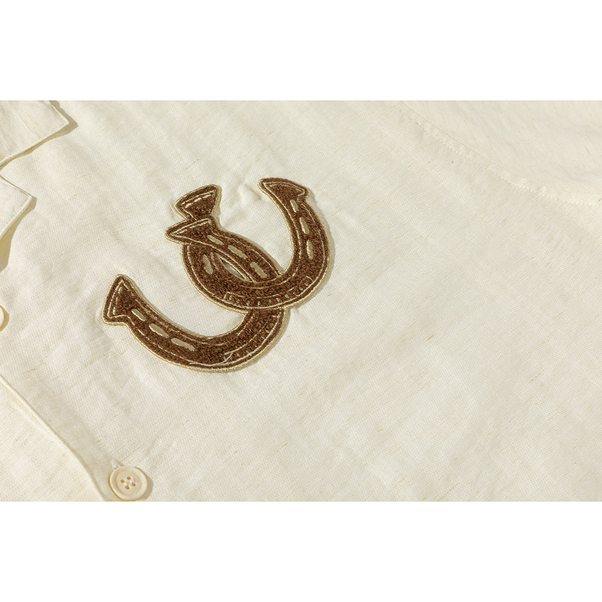 Close-up of a brown embroidered horseshoe motif on ivory shirt fabric, with a button and collar edge visible.