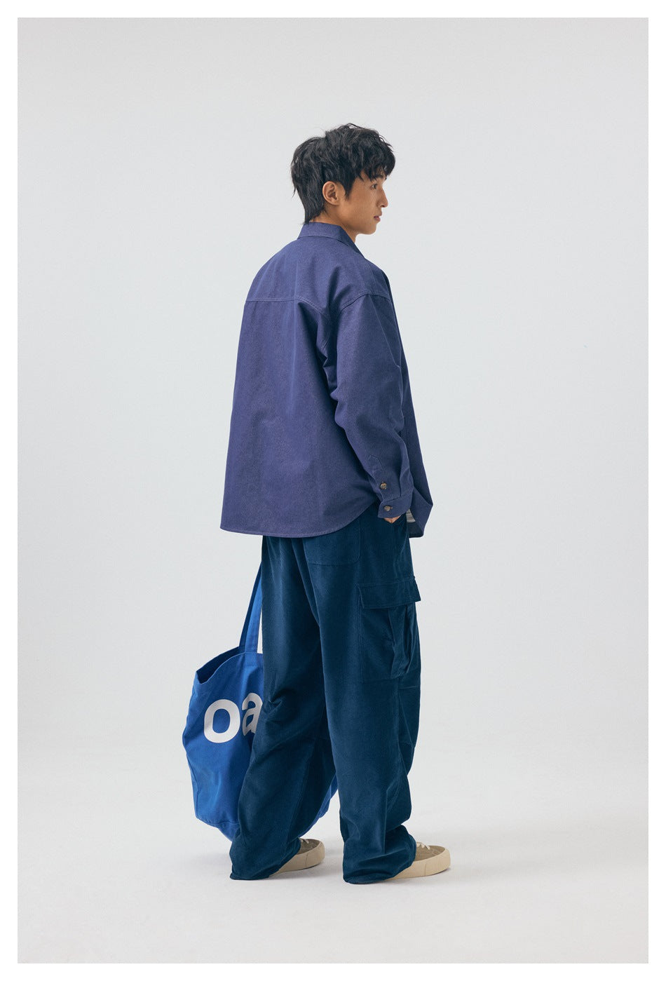 Side and rear view of a model in a blue shirt and dark cargo pants, carrying a blue tote bag.