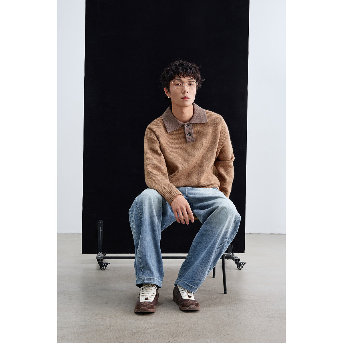 Seated model wearing a camel-brown knit polo sweater with a contrasting taupe collar and loose blue jeans.