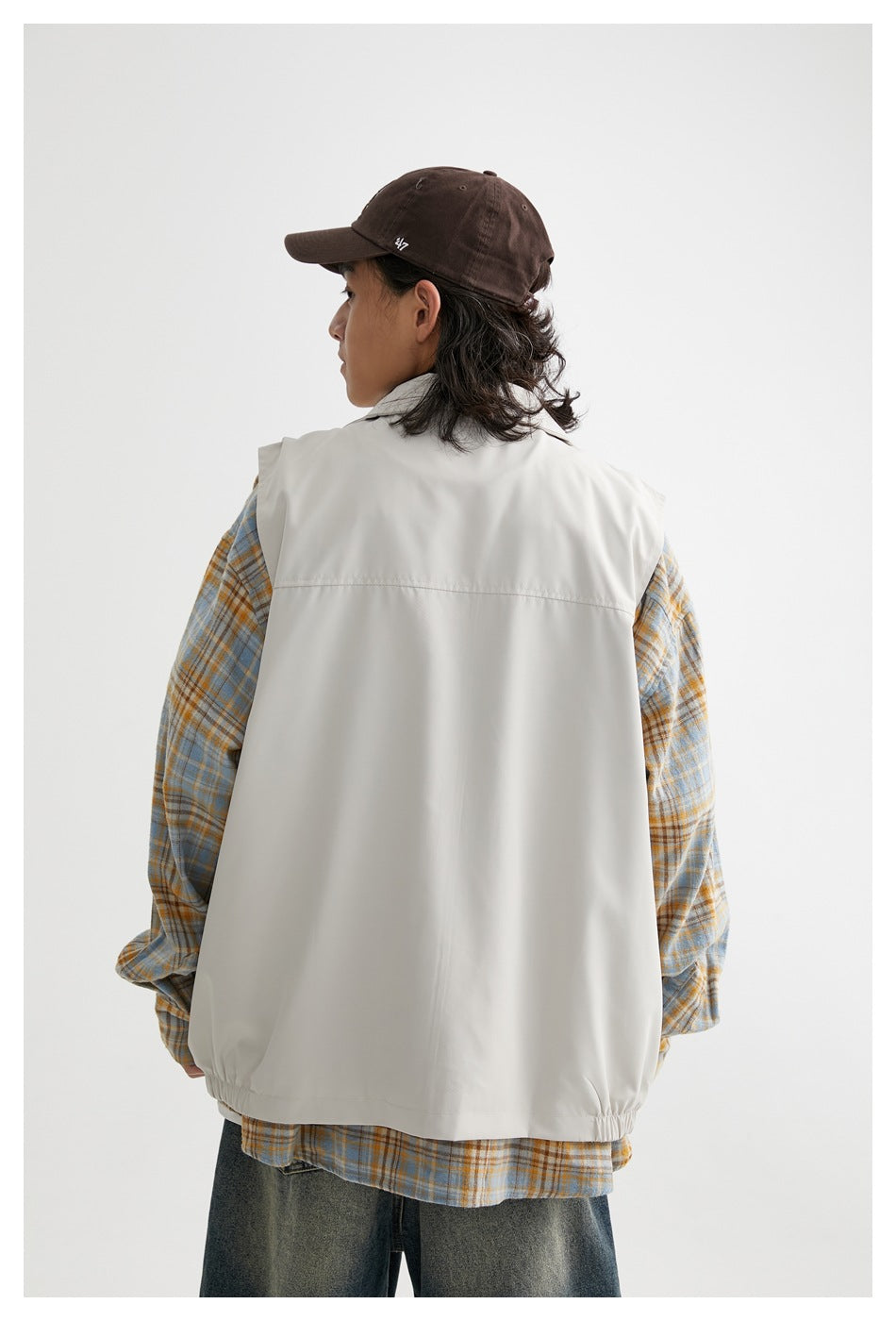 Back view of a model wearing an off-white sleeveless zip vest over a blue and ochre plaid shirt.