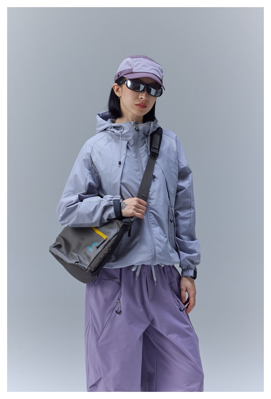 Model in a light gray zipped hooded jacket and lavender pants, shown with a crossbody bag.