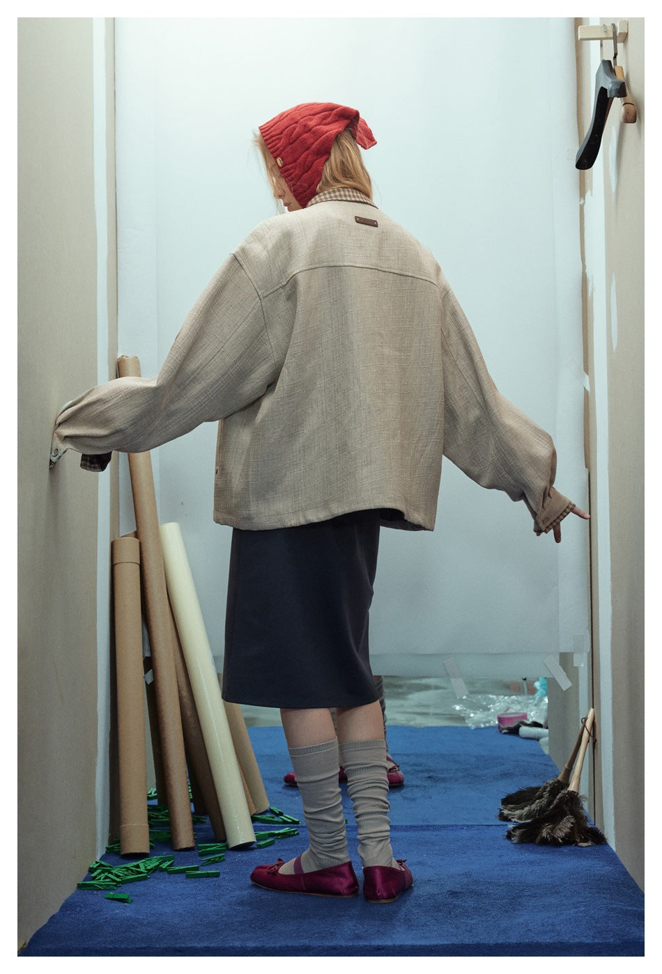 Back view of model wearing a loose beige textured jacket with back yoke seam, dark skirt and red knit hat