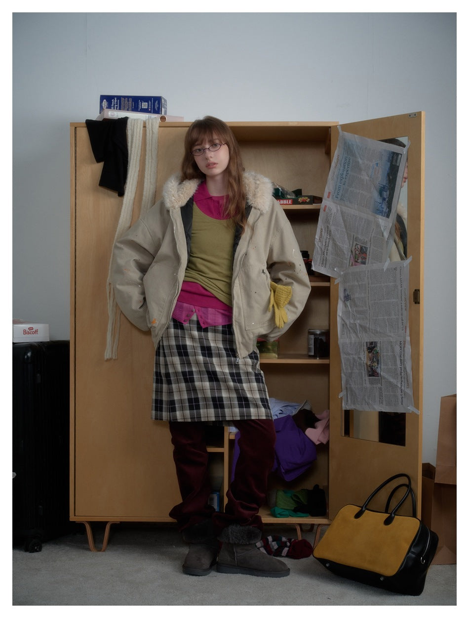 Model wearing a beige hooded zip jacket with fluffy hood trim over layered tops and a plaid skirt by a wooden wardrobe