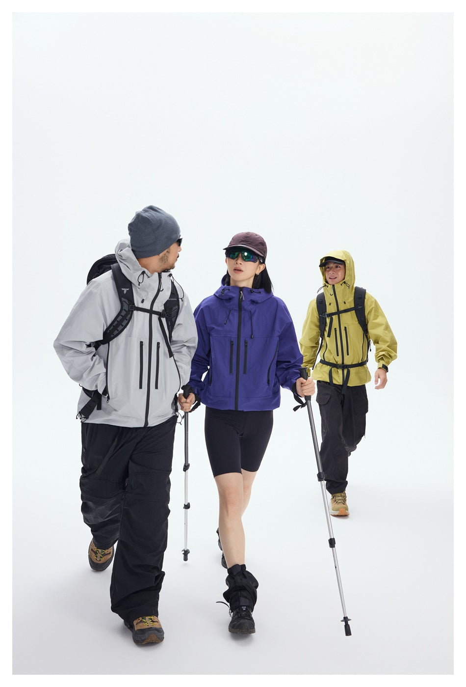 Three hikers walk in gray, purple, and yellow hooded jackets with black pants and trekking poles.