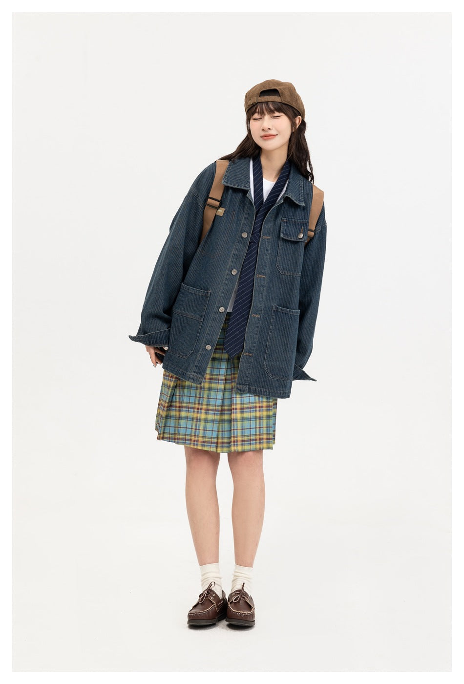 Model wearing a dark blue denim chore jacket over a plaid skirt, with a striped tie and backpack.