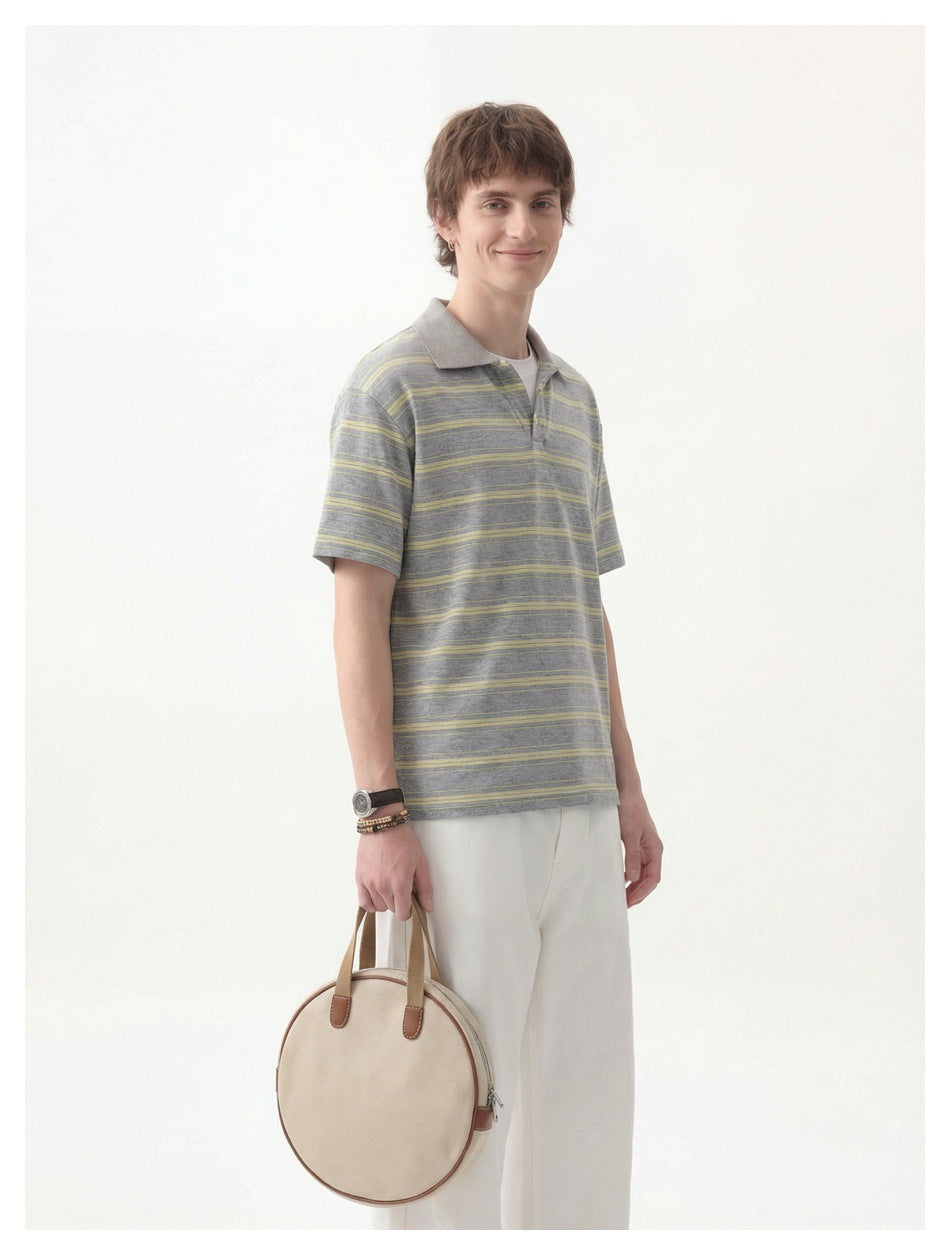 Model wearing a gray polo with pale yellow stripes and white trousers, holding a round beige bag.