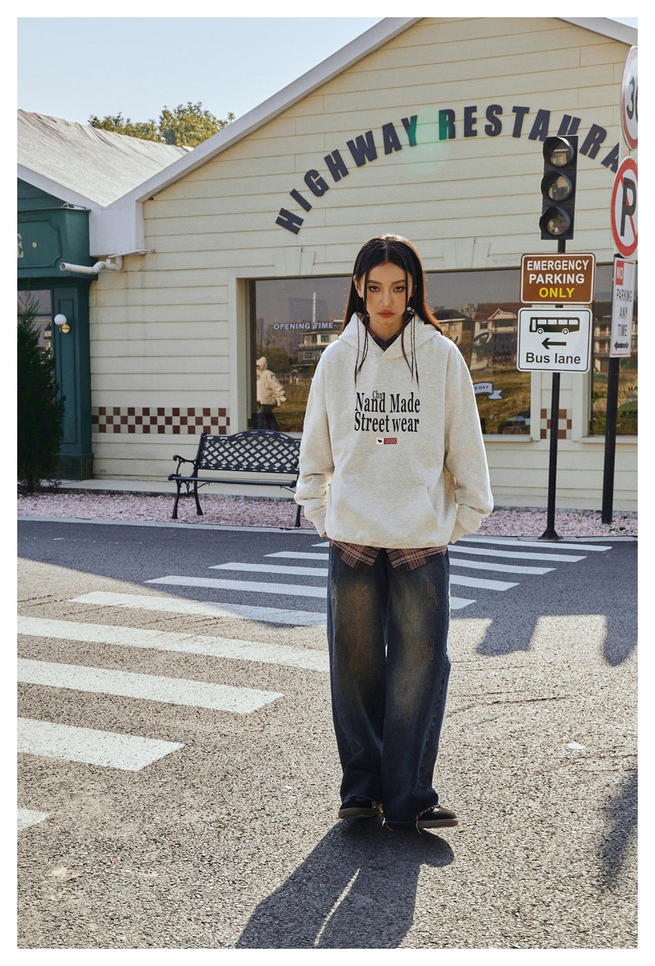 Full-length view of a model wearing a light gray graphic hoodie and loose dark jeans at a street crossing.
