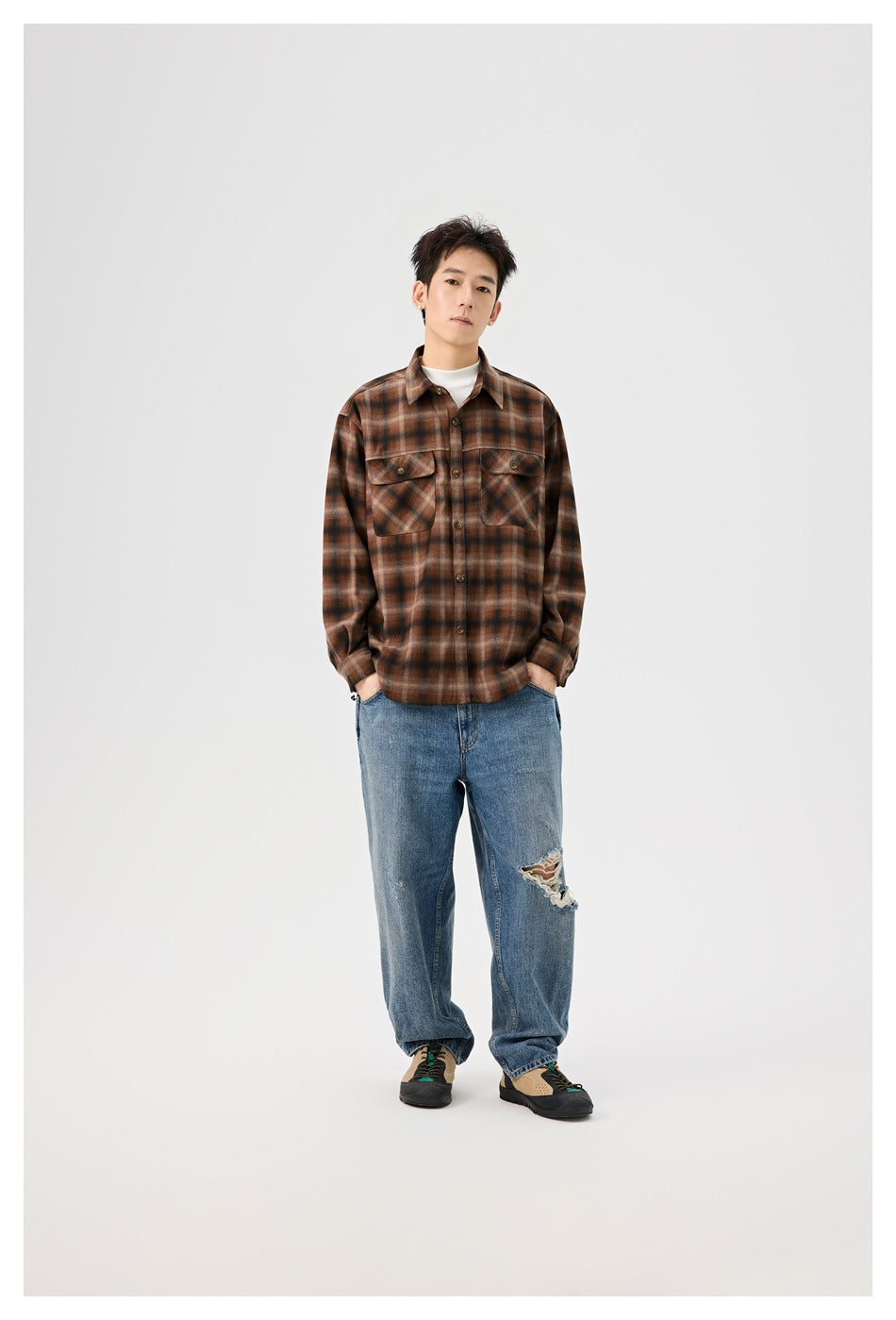 Full-length front view of a model wearing a brown plaid overshirt, white top, loose blue jeans with a torn knee, and