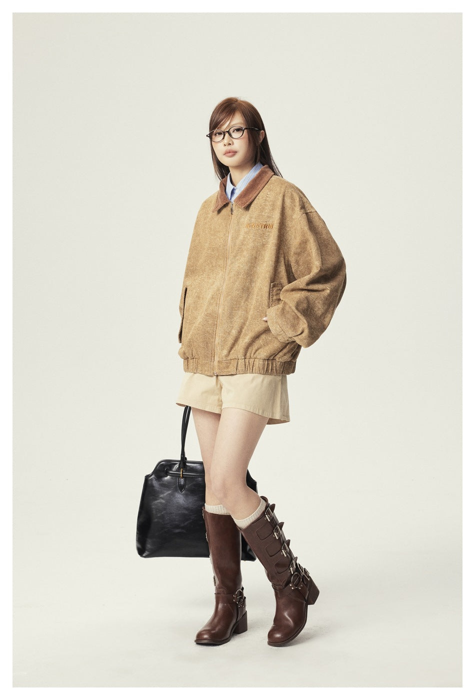 Full-length view of a model in the khaki zip jacket with beige shorts, brown buckled boots and a black tote bag