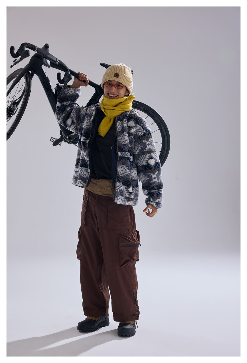 Model carries a bicycle while wearing a blue-gray and white geometric fleece jacket, yellow scarf, and beige beanie.