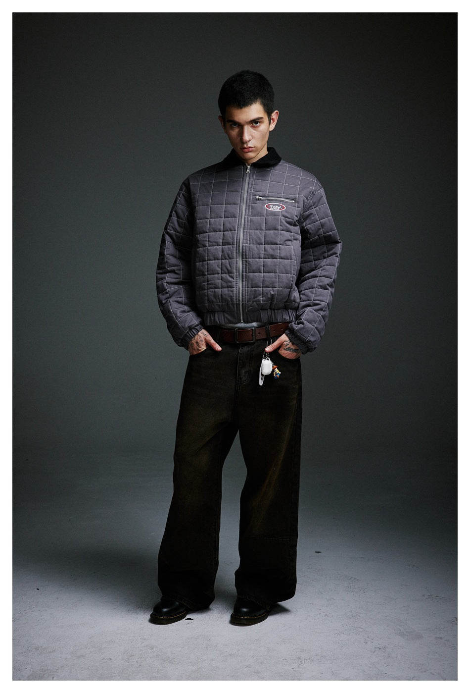 Model wearing a zipped charcoal-gray quilted jacket with a black collar, elastic hem, and zip chest pocket.