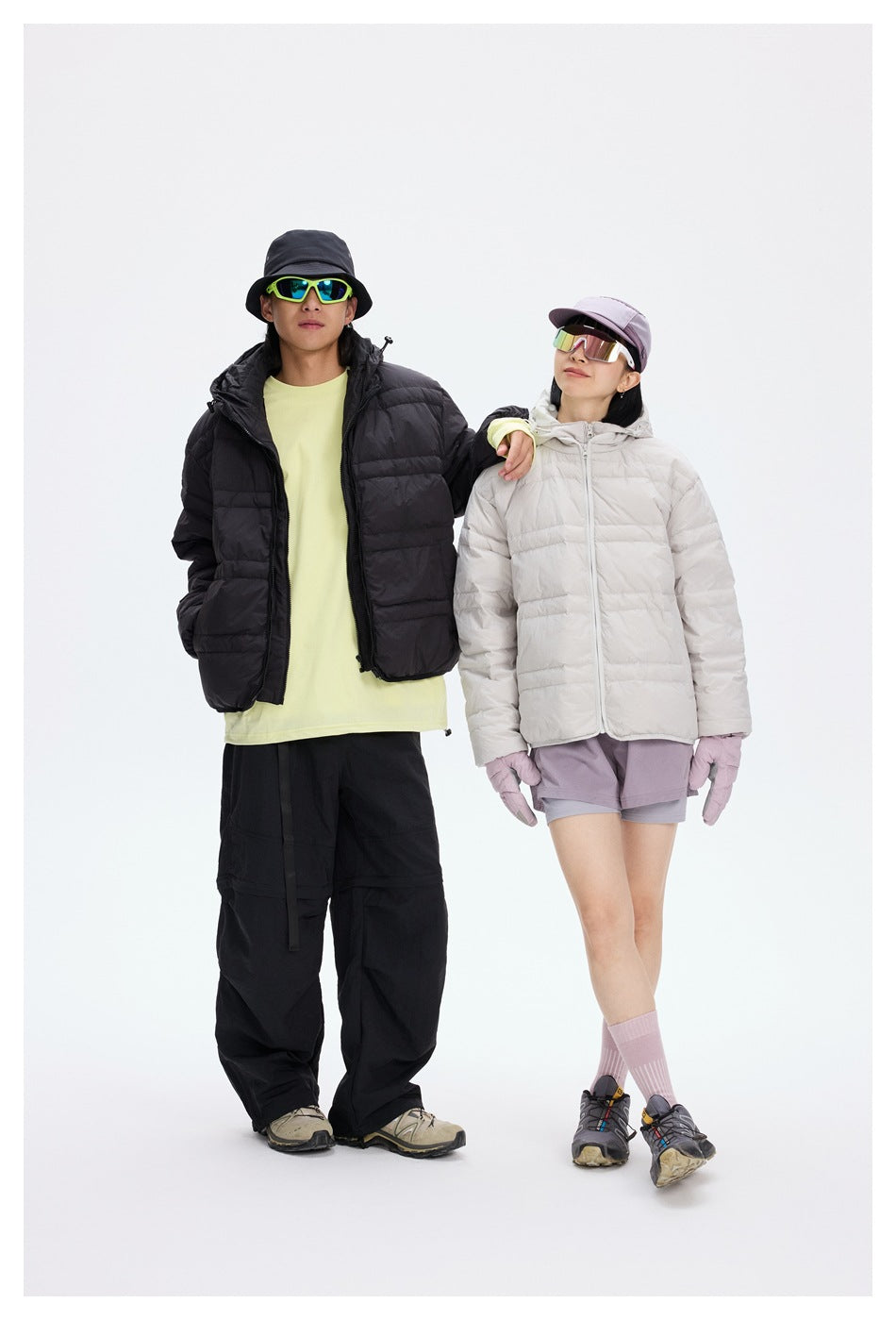 Two models pose in black and light gray quilted hooded jackets; the woman wears lilac shorts and gloves.