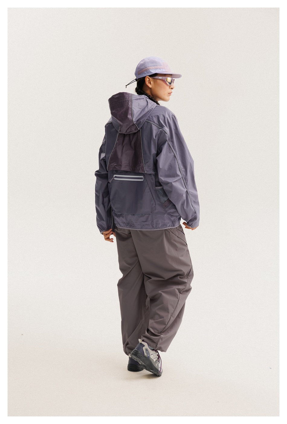 Rear view of a model wearing a purple hooded windbreaker and loose gray-purple pants.