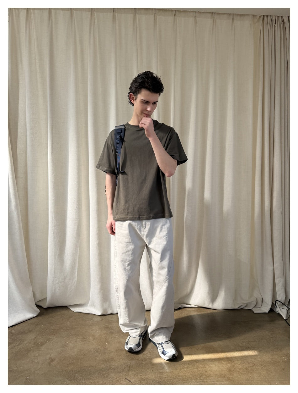 Model wearing a dark olive short-sleeve T-shirt with a small chest graphic and loose white pants.