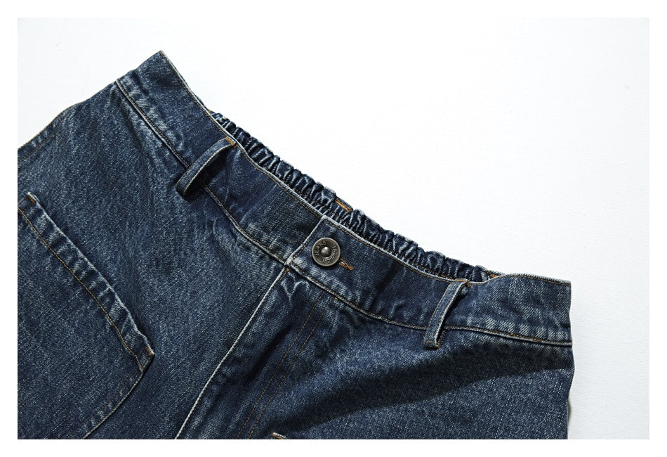 Close-up of dark blue washed denim trousers showing the gathered waistband, belt loops, and metal button.