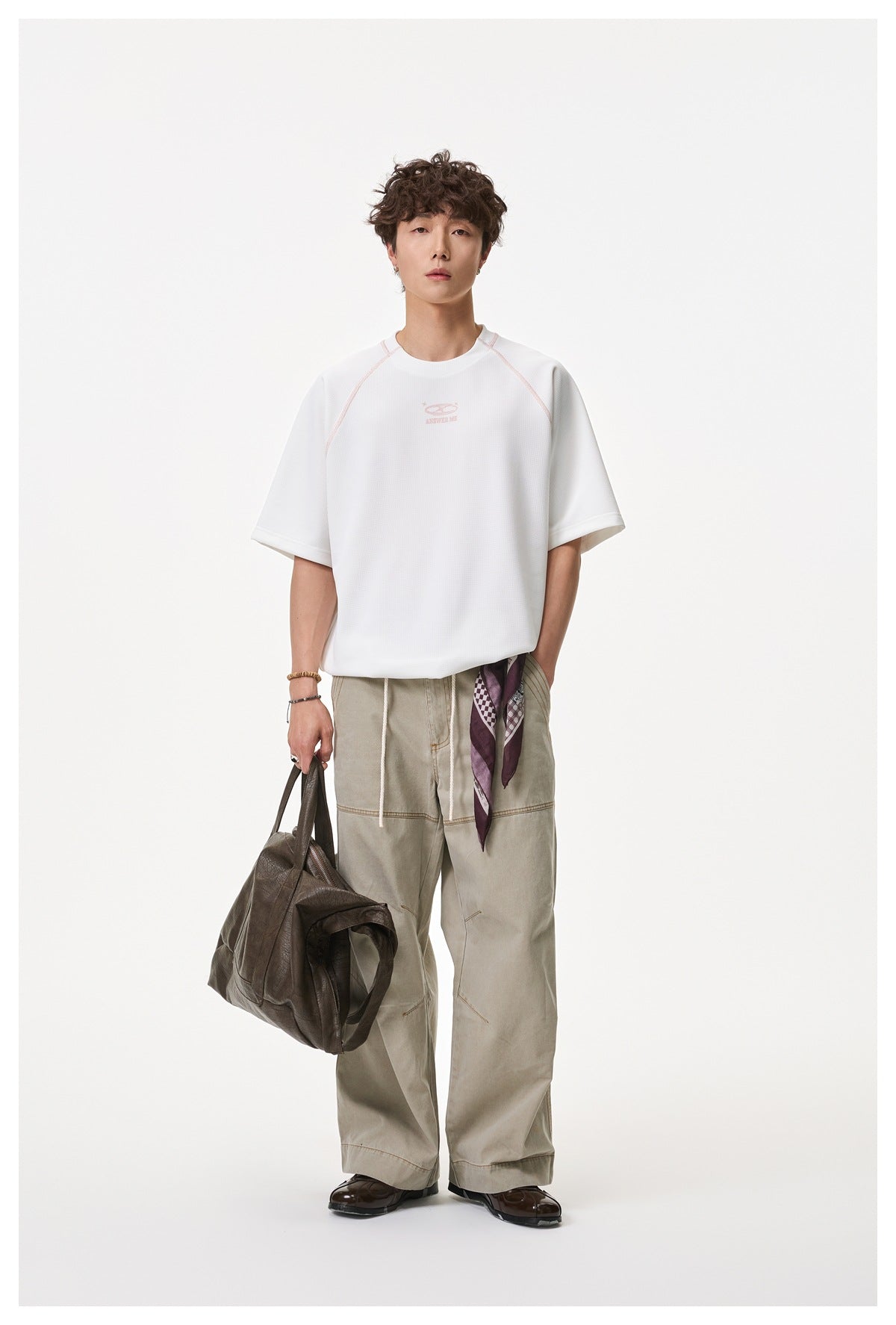 Man wearing a white waffle-textured raglan T-shirt with khaki wide-leg pants, scarf and brown leather bag, full-length