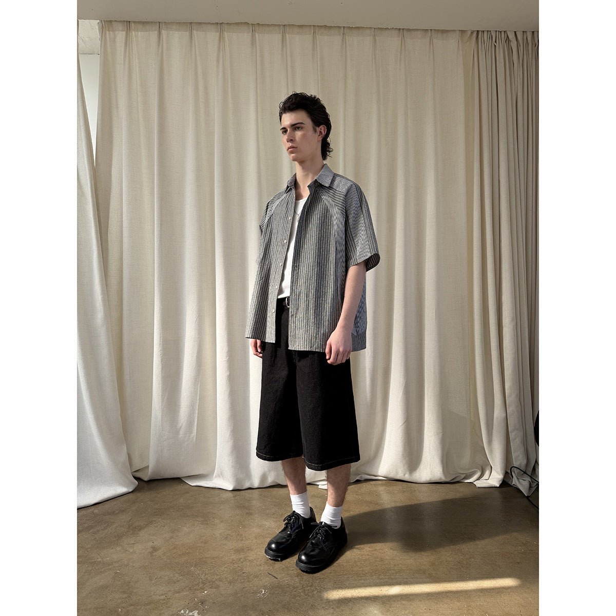 Full-length model wearing an open gray striped short-sleeve shirt over a white top and black shorts.