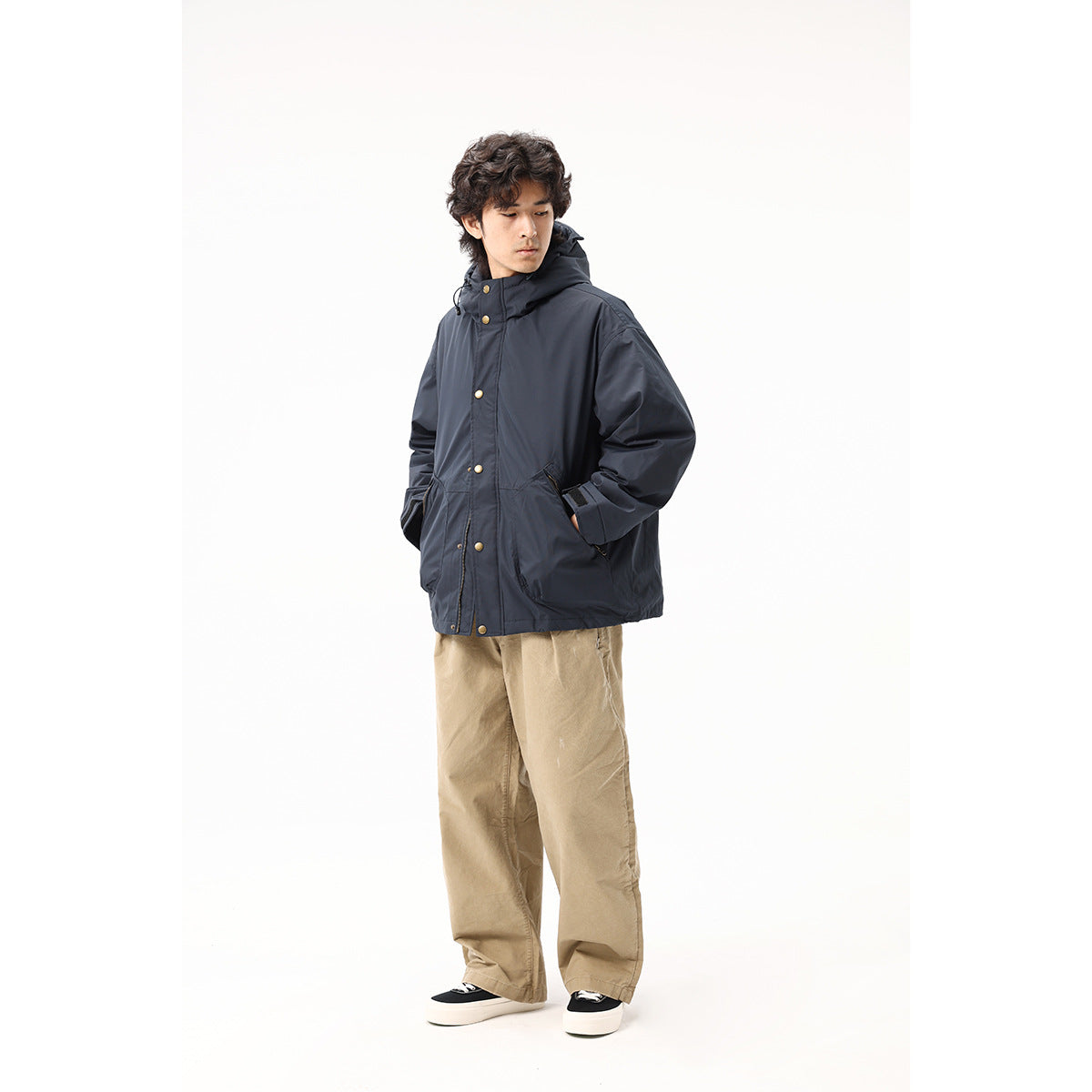 Model wearing a dark navy hooded jacket with brass-tone snaps and large pockets, paired with tan trousers.