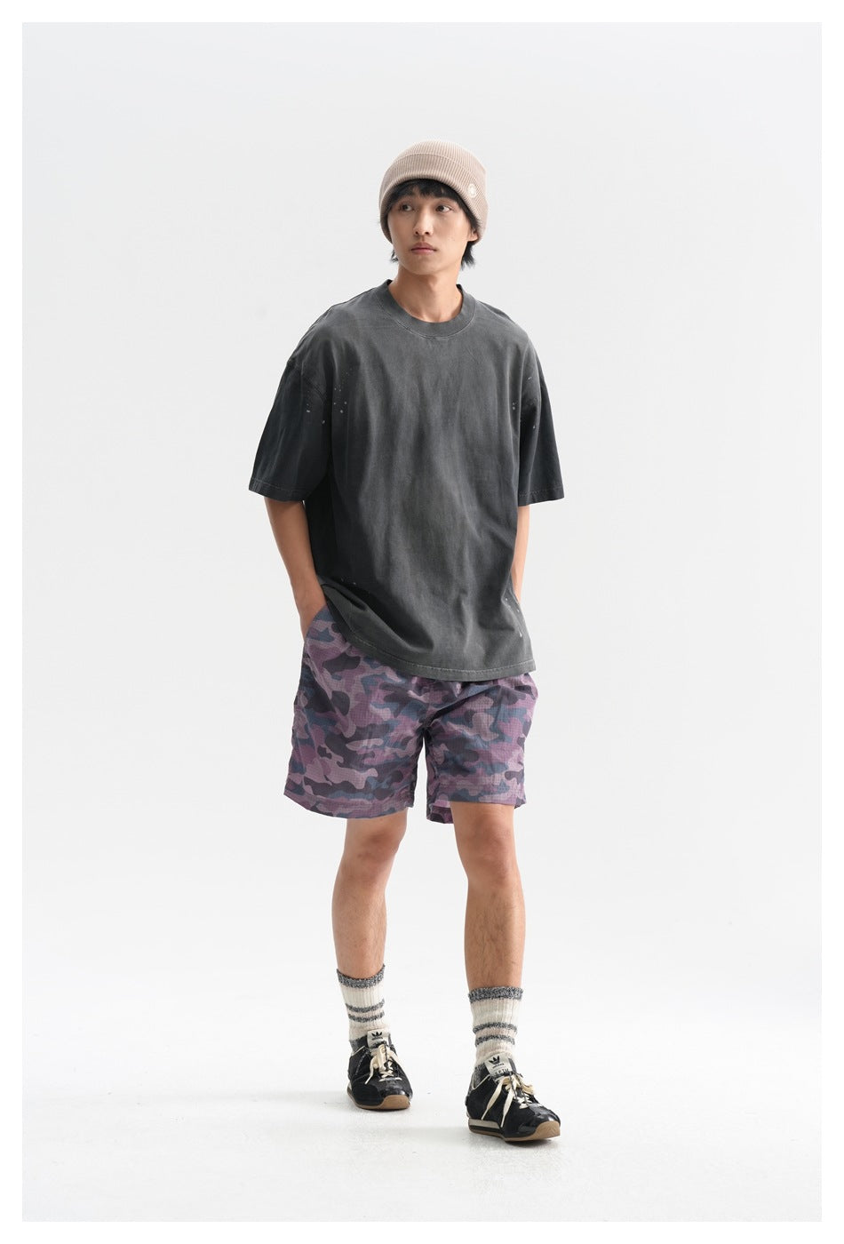 Front view of a model wearing an oversized charcoal T-shirt and purple camouflage shorts with a beige beanie.