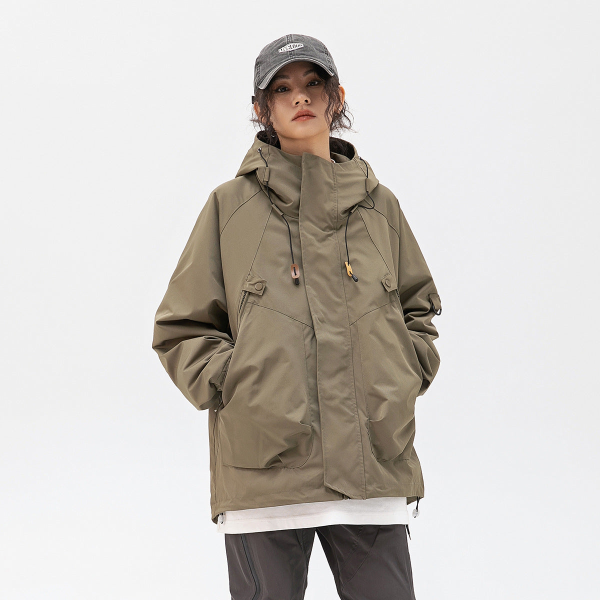 Model wearing an olive hooded jacket with a high collar, front storm flap, drawcords, and large pockets.
