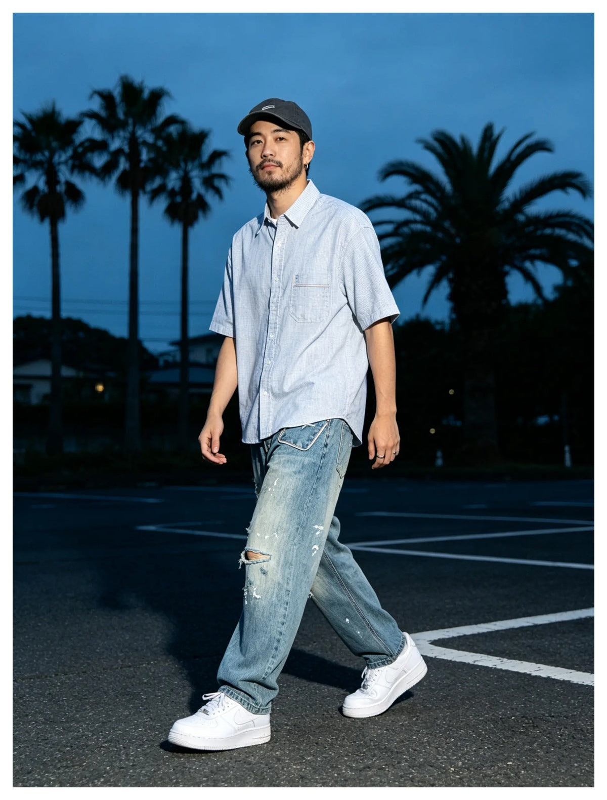 Man walking outdoors in faded blue ripped wide-leg jeans with a light blue short-sleeve shirt and white sneakers