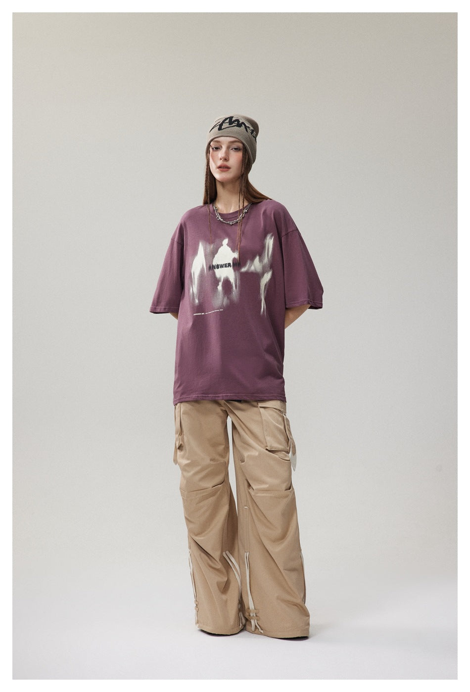 Model wearing a purple oversized T-shirt with blurred white figure graphic, beanie and wide-leg beige cargo pants