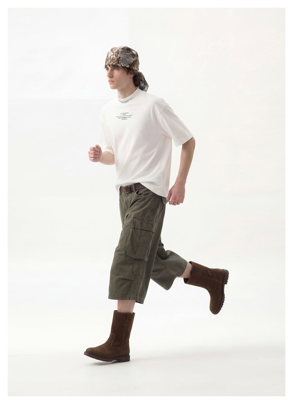 Model in profile wearing a white printed T-shirt, olive cargo trousers, brown boots, and a camouflage headscarf.
