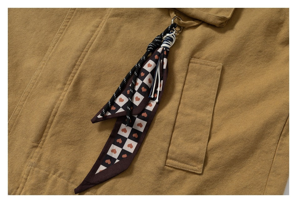 Close-up of a mustard-brown jacket with a rectangular pocket and a dangling black-and-white heart-patterned fabric accessory.