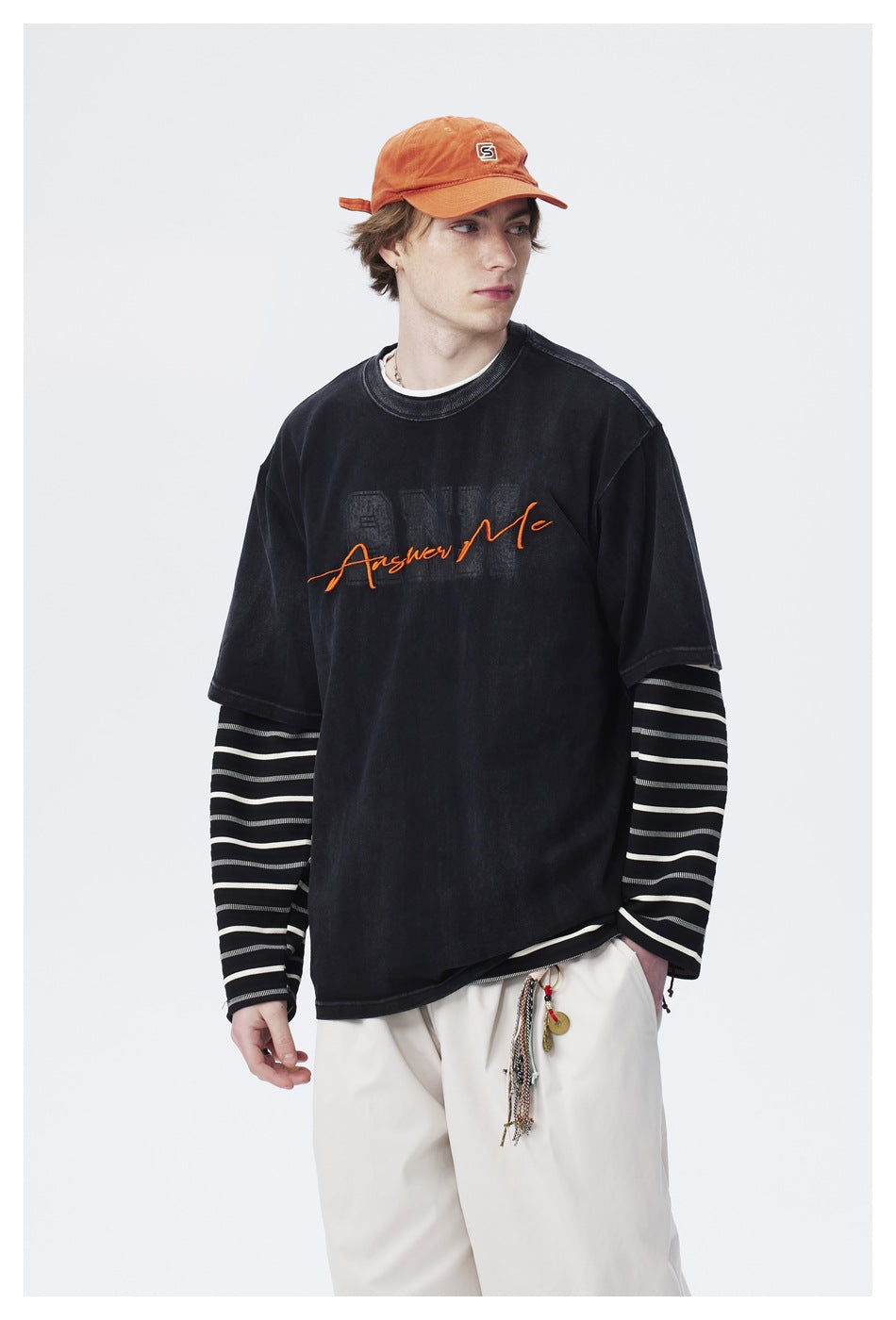 Closer front view of a model in a black T-shirt with orange script layered over striped sleeves and loose white pants.