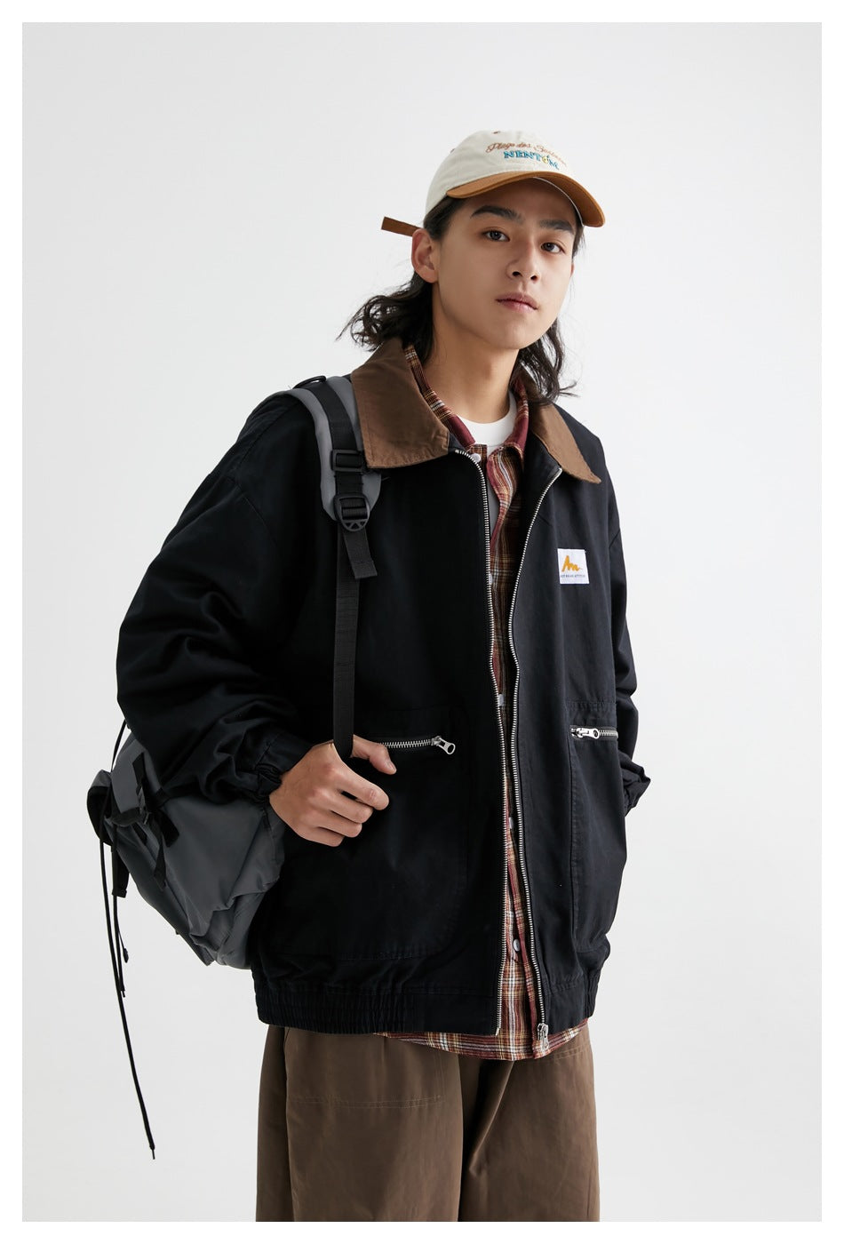 Model wearing a black zip jacket with a brown collar and zip pockets, shown with a backpack and brown trousers.