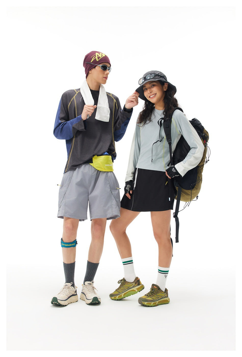 Two full-length models in coordinated hiking outfits, wearing long-sleeve tops, shorts, hats, and backpacks against a white