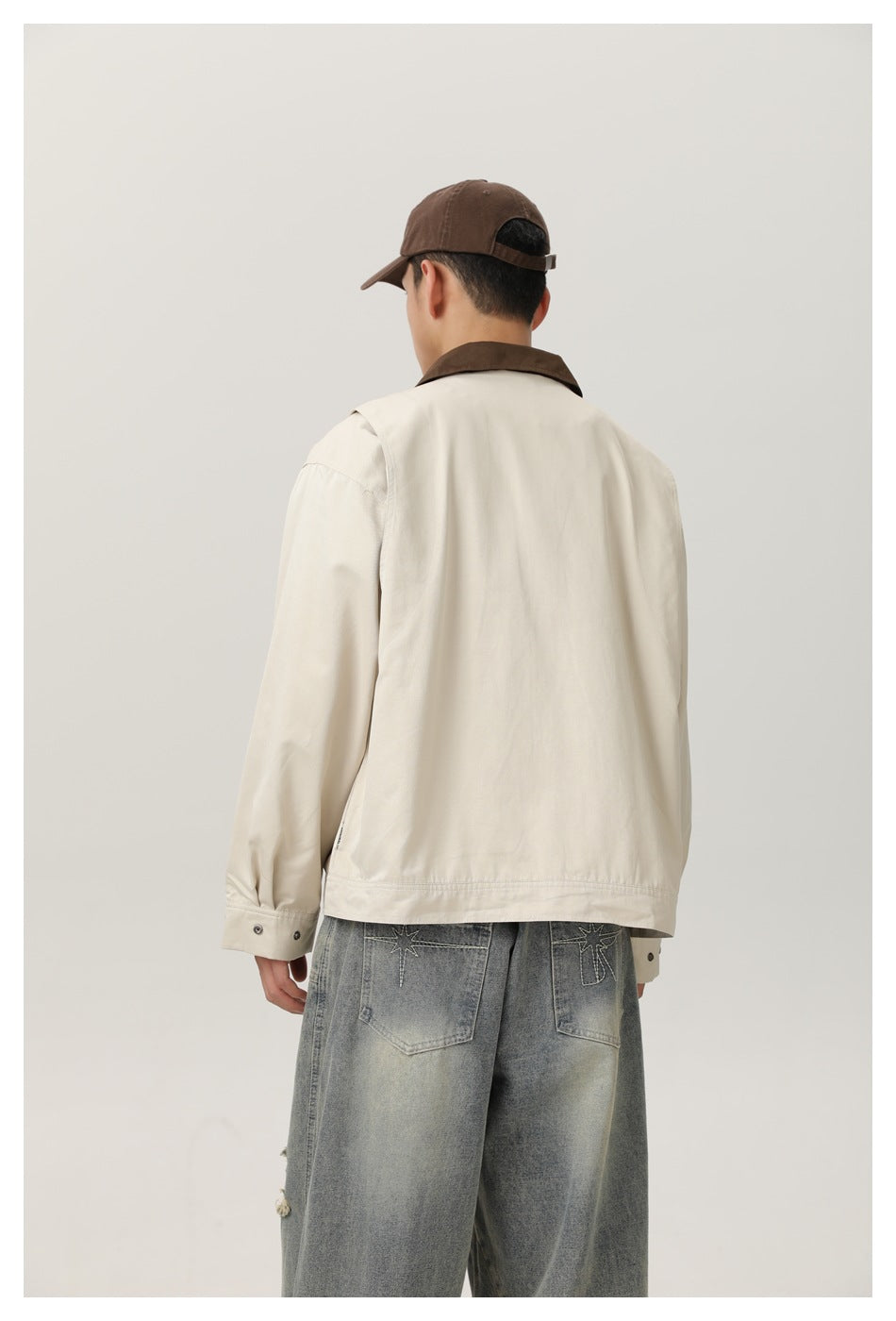Back view of model wearing beige jacket with brown collar, brown cap and faded embroidered jeans