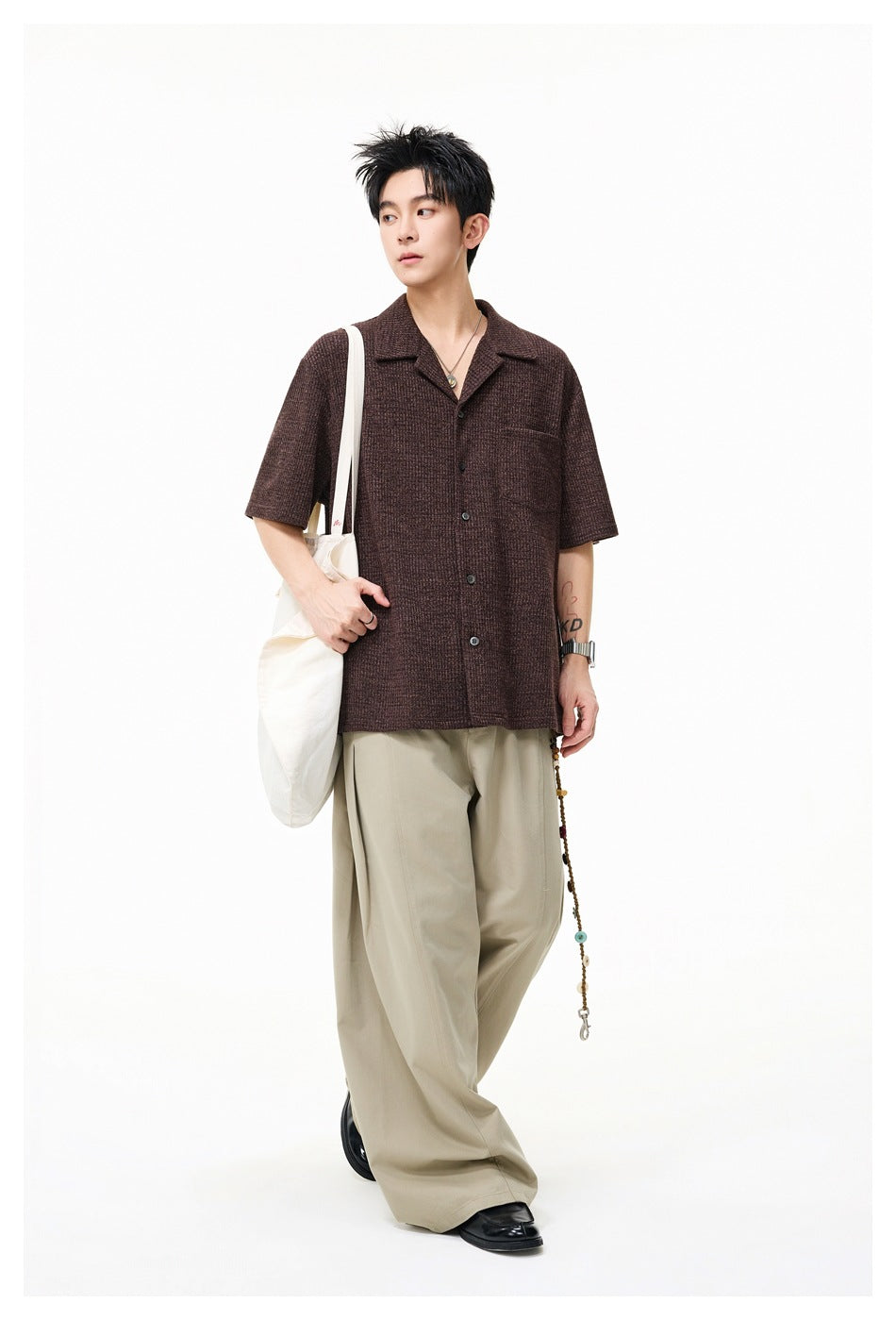 Full-length model wearing a brown short-sleeve button-up shirt with a notched collar and chest pocket, carrying a canvas